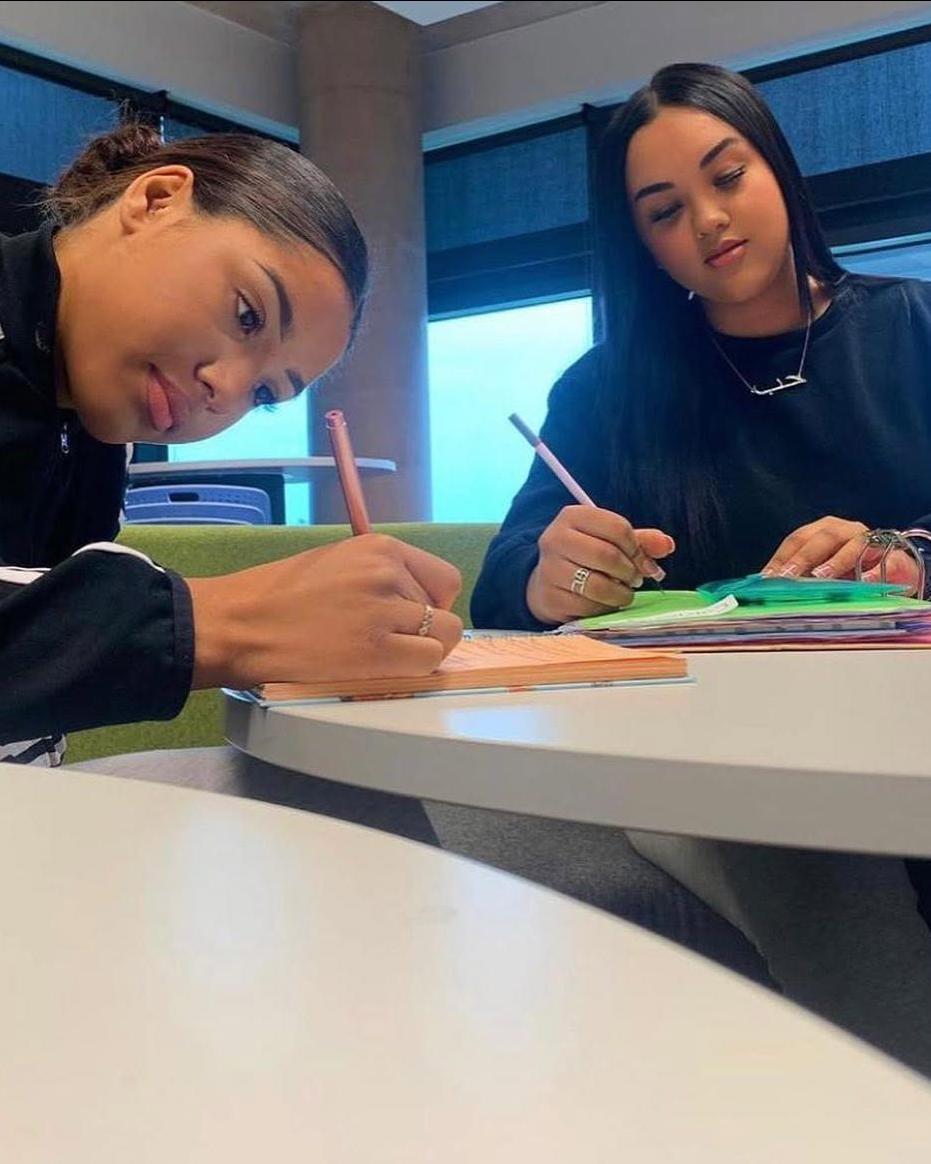 2 girls sat together in a table; writing something in their notebooks