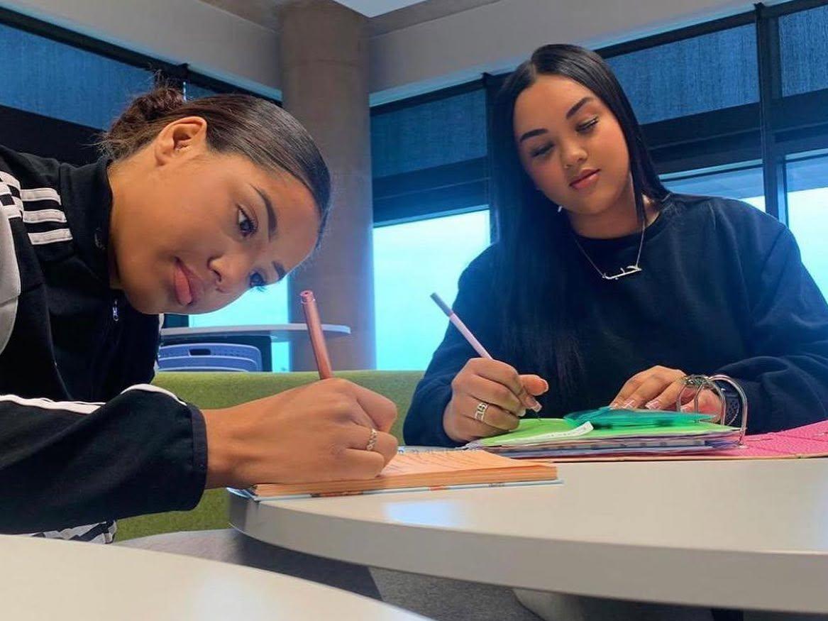 2 girls sat together in a table; writing something in their notebooks